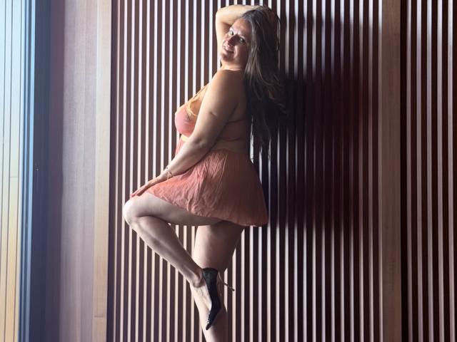 BadAngel with big tits, thick ass, and long brown hair, wearing a pink dress, black heels, and looking over shoulder, standing against a wooden slat wall.