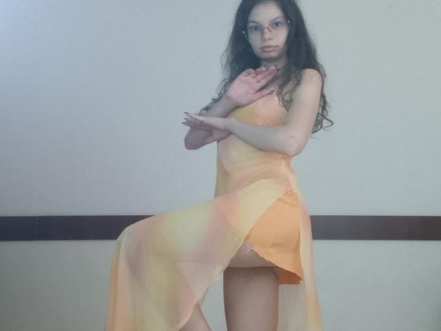 VikkiRi, com cabelos longos e escuros, óculos e pele clara, vestindo um vestido laranja transparente, cobrindo seus pequenos seios com uma mão, em pé com uma perna levantada, expondo sua vulva..