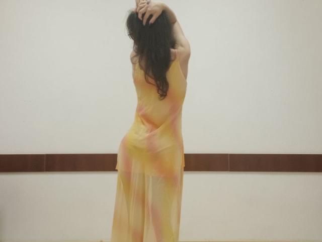 VikkiRi está de espaldas a la cámara, con los brazos levantados, sosteniendo su largo cabello oscuro. Lleva un vestido amarillo, con un tono rosado, semitransparente que revela sus glúteos. Habitación sencilla, de color beige..