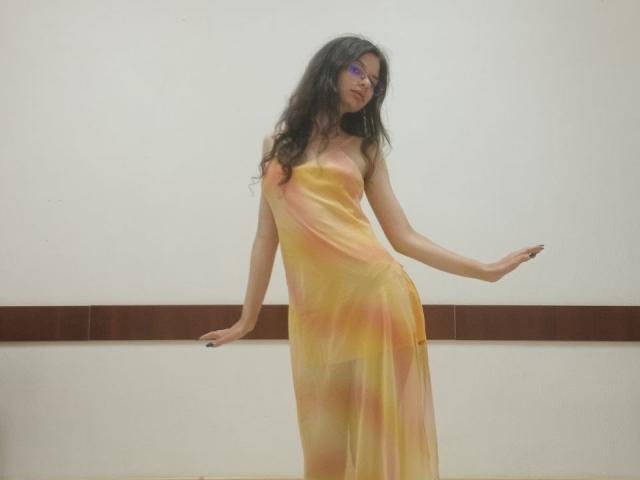 VikkiRi, avec de longs cheveux noirs, portant une robe jaune, semi-transparente, debout, bras droit levé, bras gauche reposant sur une bande horizontale marron, portant des lunettes violettes..