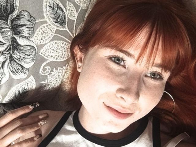 Kasandra Grothe, met rood haar, sproeten en oorringen, lachend naar de camera, draagt een zwart-wit top, ligt neer en heeft een hand met glinsterende nagels in de buurt van haar gezicht..