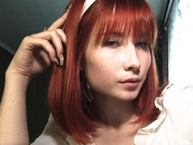 Kasandra Grothe, con i capelli rossi, una fascia bianca in testa e la frangia, che si tiene i capelli, indossa una camicia bianca e guarda leggermente di lato, con un'espressione neutra..