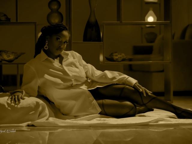MidnightSins is a dark-skinned woman with medium-sized tits, lying on a bed in a yellow-brown lit room. She's wearing a white shirt, black pantyhose, and earrings.