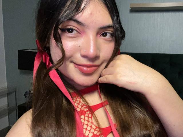 LunaRosseX, usando lingerie de rede vermelha, fitas rosa no cabelo castanho, com um piercing no nariz e um sorriso adorável, apoiando o queixo na mão. Seus seios estão parcialmente visíveis..