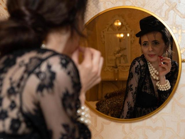KamiLove in black lace dress, pearl necklace, and black hat, looking at round mirror, touching red lipstick. Reflection shows her cute face, focused expression, and elegant style.