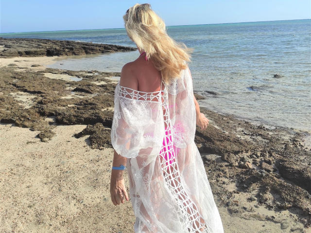 Sofie: Sexo quente com cabelo loiro, vestindo uma peça branca de renda, mostrando um biquíni rosa, em pé em uma praia rochosa, com o oceano ao fundo, luz do sol, pulseira azul, de costas para a câmera..