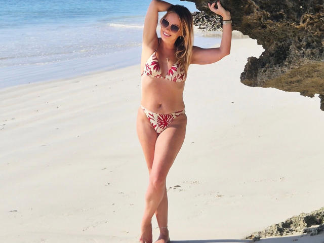 Sofie : Sexe torride dans un bikini rouge à motifs floraux, debout sur une plage, les bras au-dessus de la tête, portant des lunettes de soleil, regardant l'appareil photo, rocher sur la droite, eau bleu clair, sable blanc..
