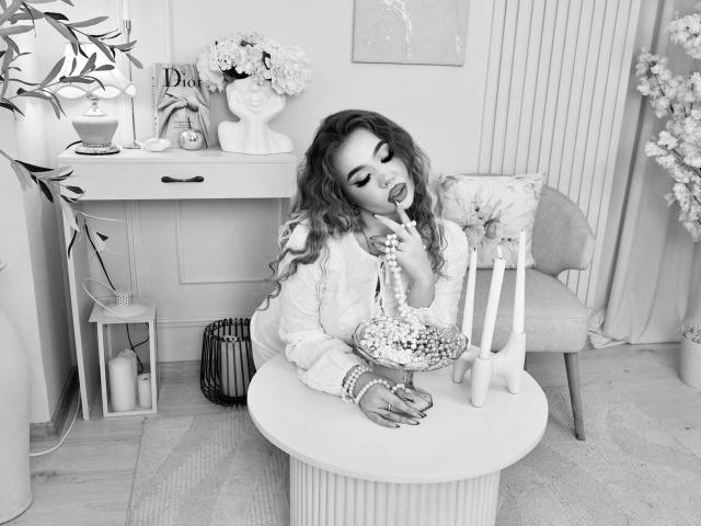 Stacy Yellig, com cabelos ondulados, vestindo uma blusa branca, chupando o dedo, segurando um cachimbo decorado com miçangas e um pequeno unicórnio branco, sentada em uma mesa redonda em um quarto aconchegante..