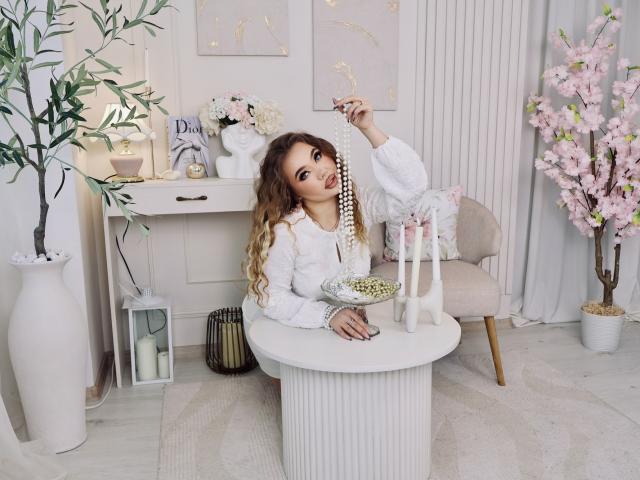 Stacy Yellig, com cabelos loiros cacheados, vestindo um suéter branco e fofo, segurando um colar de pérolas, com gotas de chuva no rosto, sentada em uma mesa branca, com velas cor-de-rosa, um vaso branco e uma árvore de cerejeiras cor-de-rosa..