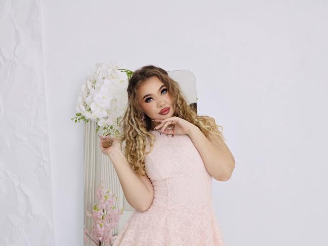 Stacy Yellig, com cabelos loiros cacheados, vestindo um vestido rosa de renda, tem a mão perto da boca e está em pé ao lado de uma parede branca com flores. Ela tem um rosto adorável..