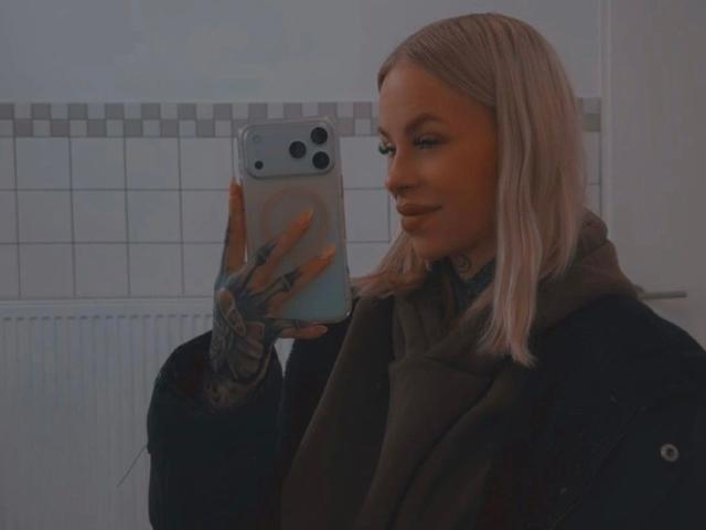 Angelxxx1-hot with blonde hair, tattoos on hand, green scarf, black jacket, holding phone, taking mirror selfie, slight smile, visible nose, subtle makeup.