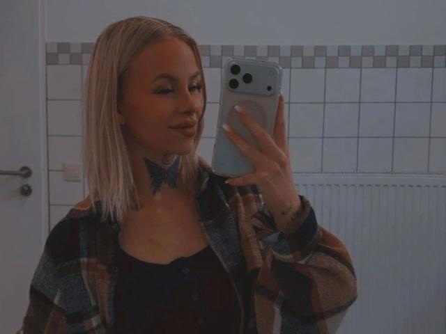 Angelxxx1-hot with blonde hair, wearing a plaid shirt, black top, and blue bowtie, taking a mirror selfie with an iPhone. Her tits are small, she's smiling.