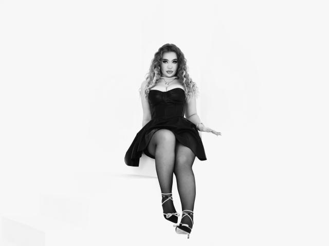 Stacy Yellig a les cheveux blonds ondulés, porte une robe noire sans bretelles et est assise les jambes croisées, avec des talons hauts, montrant sa poitrine et ses fesses, sur un fond blanc..