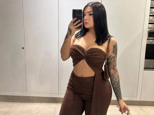 KarolGoddes con un top marrón sin tirantes que muestra sus grandes senos, pantalones marrones de cintura alta, cabello negro, tatuajes en el brazo derecho, sosteniendo un teléfono, de pie en la cocina, selfie frente al espejo..