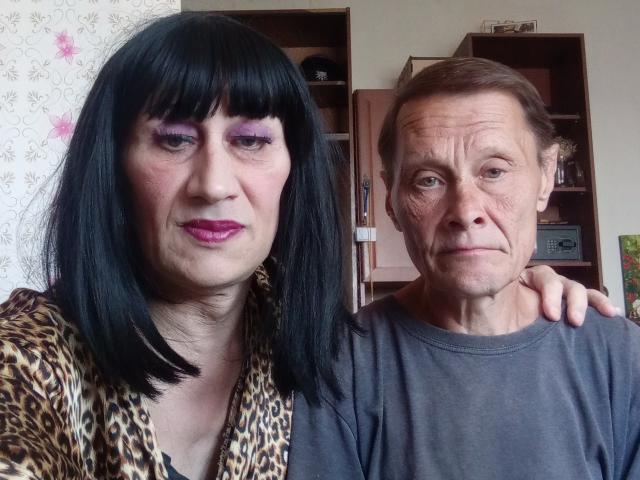 ArtemidaImax, met zwart haar, paarse oogschaduw, een top met luipaardprint en roze lippenstift, staat naast een oudere man met een grijze trui en een vermoeide uitdrukking, zijn hand op haar schouder..