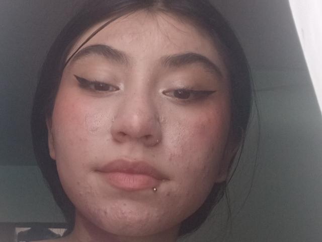 Vallee's Gesicht, mit schwarzem Eyeliner, einem Nasenring und einem Piercing in der Unterlippe, zeigt leichte Akne und einen neutralen Ausdruck, dunkles Haar und einen leicht geröteten Teint..
