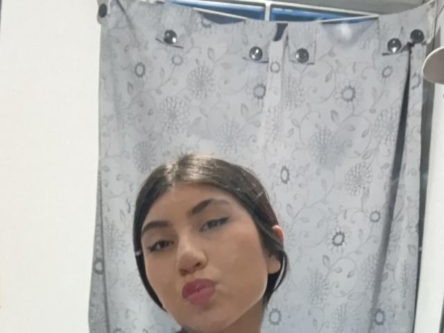 Vallee con labbra rosse e piene, capelli castano scuro e un trucco leggero, viso con le labbra strette in un selfie, sfondo con tenda floreale bianco-grigia, parte superiore di una tenda blu visibile..