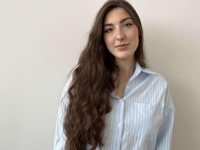 Tia Brounson, com longos cabelos castanhos ondulados, vestindo uma camisa azul listrada com botões, em pé em frente a uma parede branca, sorrindo suavemente, nariz pequeno, pele clara, olhos verdes..