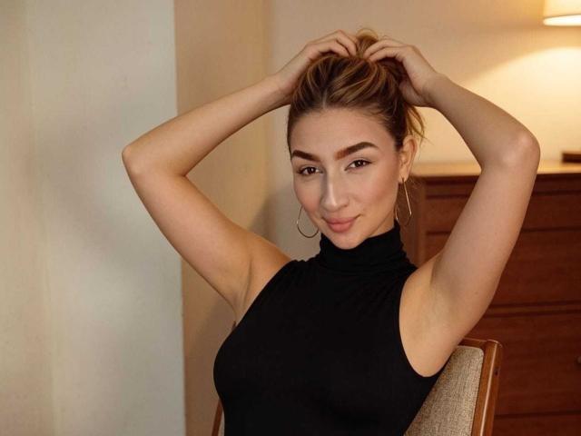 Abby, com as mãos no cabelo castanho, vestindo um cachecol preto sem mangas, mostrando os seios, usando pequenas argolas nos ouvidos e com um leve sorriso, sentada em uma cadeira de madeira..