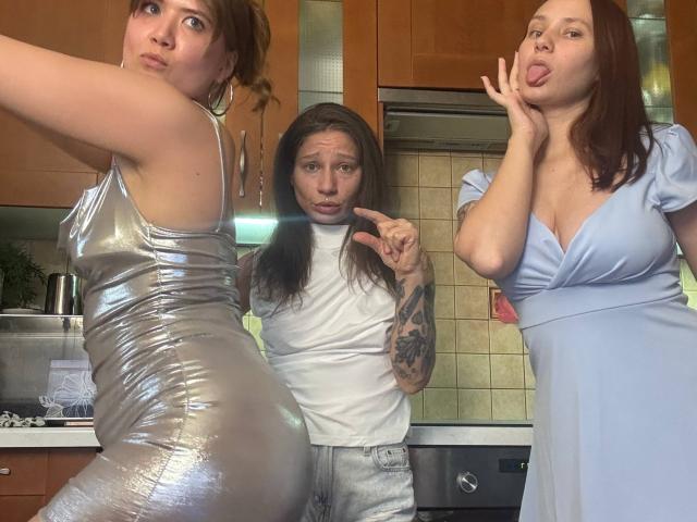 Three HotCaramelsTrio in a kitchen. One with silver shiny dress and big ass, one with white t-shirt and tattooed arm, one with blue dress and tongue out.