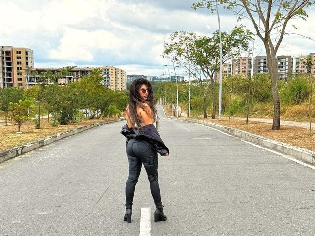 AntonelaRiveiro se tient sur une route, dos à la caméra, nue sur le torse, avec un pantalon noir et des bottes, ses fesses bien visibles, de longs cheveux noirs, un ciel nuageux, des arbres et des bâtiments en arrière-plan..