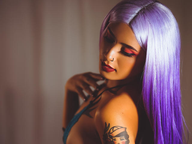 VelvetMusee, com cabelo roxo-prateado, pálpebras vermelhas, piercing no nariz e braço tatuado, exibindo pequenos seios em uma blusa azul, com as mãos perto do peito, olhos fechados..