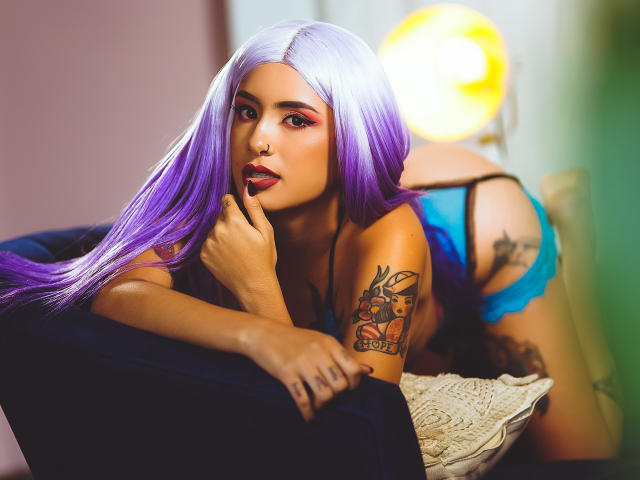 VelvetMusee com cabelo roxo, lingerie de renda azul, braço tatuado, dedo na boca, deitada em um sofá escuro, luz amarela ao fundo, sexy, sedutora, cabelo comprido, sombra vermelha..