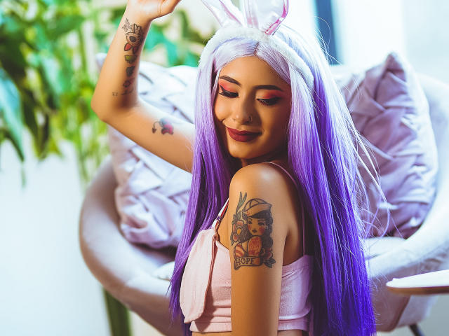 VelvetMusee con capelli viola, orecchie da coniglio rosa e trucco, che mostra i suoi tatuaggi, sorridendo, con un braccio alzato, indossa una maglietta rosa, seduta su una sedia rosa, mettendo in mostra il suo seno..