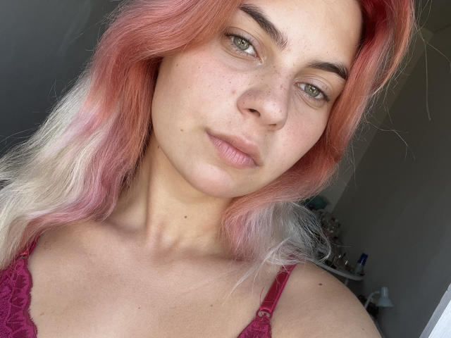 SadeSeney, avec ses cheveux aux reflets roses-blancs, son visage constellé de taches de rousseur, ses yeux verts et son haut en dentelle rose, a l'air légèrement ennuyée, avec les lèvres pincées. Sa poitrine n'est pas visible. Lumière douce. Autoportrait décontracté..