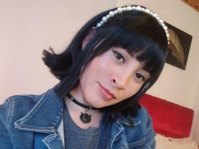 IssisDiosa has short black hair, a pearl headband, black eyeliner, a blue denim jacket, and a black choker with a pentagram. She's smiling slightly with pink lipstick.