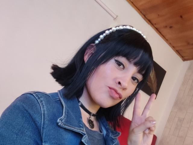 IssisDiosa with short black hair, pearl headband, blue denim jacket, black choker, making a peace sign, pouty lips, and light skin, in a selfie.