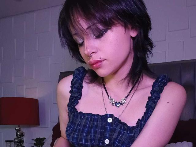 ViolettClay with short black hair, nose piercing, and heart necklace, wearing a blue plaid dress with ruffled straps, looking down with closed eyes, and slightly pouty lips.