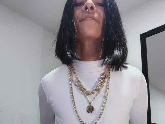 AmelieBlake has black shoulder-length hair, a nose ring, and wears a white shirt with gold chain necklaces. She's smiling down at the camera.