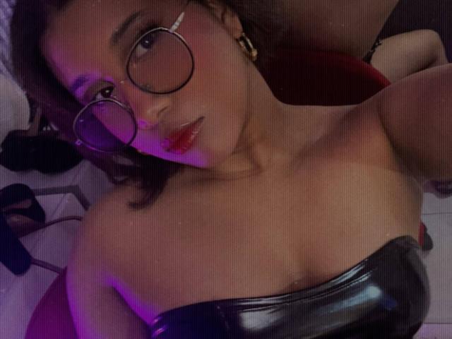 LoreDuval with glasses, red lips, and a shiny black strapless top, showing her tits, lying down, with purple lighting and a relaxed expression.