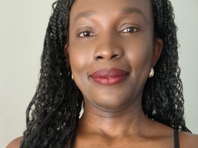 Flirtdoll-hot tiene la piel de color marrón oscuro, cabello negro rizado, lápiz labial rojo y está sonriendo, con pequeños aretes dorados. Lleva una blusa negra..