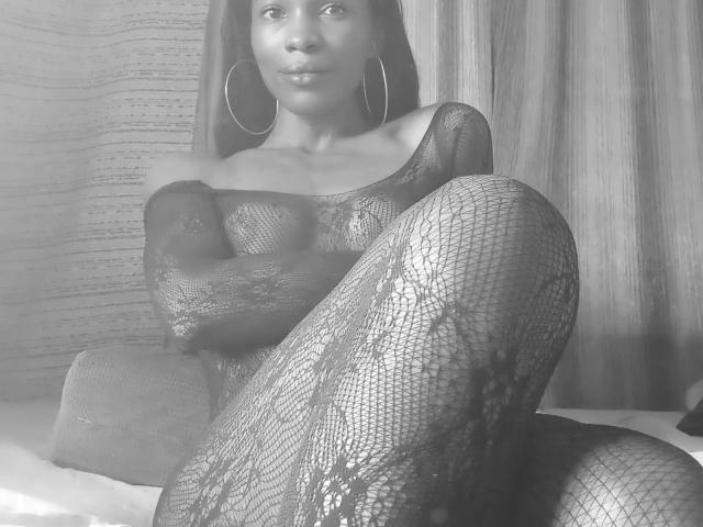 Flirtdoll, atractiva con encaje negro de rejilla, sentada con las piernas cruzadas, con grandes aros en las orejas, mirando a la cámara, fondo gris, blanco y negro, sus pechos y vulva ligeramente visibles a través del encaje..