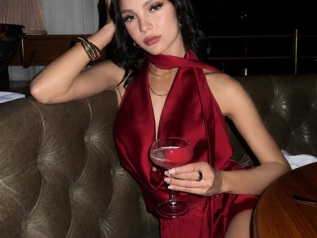 Thomasine Charlebois mit dunklem Haar, die ein rotes Seidenkleid trägt, einen roten Cocktail hält und auf einem grünen Ledersofa sitzt, mit erhobenen Arm, einem goldenen Armband und einem Ring..
