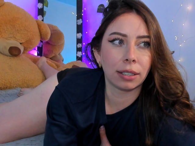 JennyLawry, mit langen braunen Haaren, blauen Augen und schwarzem Eyeliner, liegt auf dem Bauch, trägt ein schwarzes Hemd und befindet sich neben einem großen, gelben Teddybären, wobei ihr Hintern sichtbar ist..