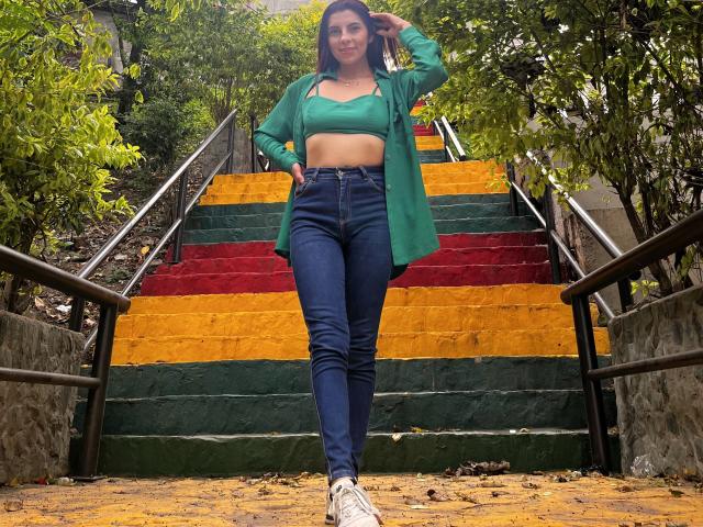 Carla Torres está de pie en unas escaleras de colores, vistiendo una camiseta verde corta, una chaqueta verde abierta, unos vaqueros azules de cintura alta y zapatillas blancas, con la mano en el pelo, mirando a la cámara..