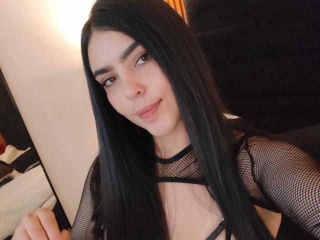 LaurynBm met lang zwart haar, die een zwarte kousenbroek top draagt, waarbij haar borsten zichtbaar zijn, met een lichte glimlach, bruine ogen, lichte huid en een subtiele make-up, in een gezellige omgeving..