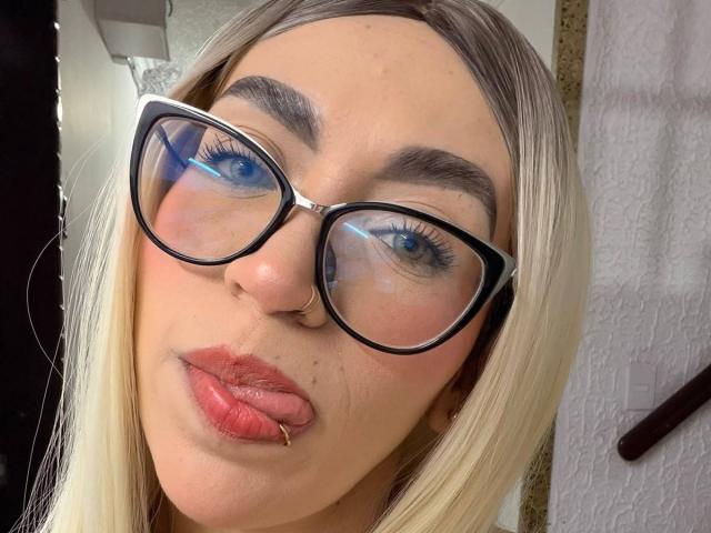 CamilleJuicyHot has blonde hair, blue eyes, thick black glasses, a nose ring, and is sticking out her tongue with pink lipstick. Her eyebrows are dark and well-defined.