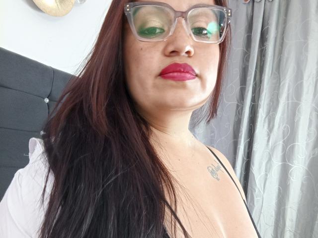 GinebraVargas with long dark brown hair, green glasses, red lipstick, small tattoo on chest, black spaghetti strap top, showing her tits, standing in front of gray curtains.