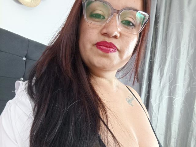 Ginebra Vargas, con rossetto rosso, ombretto verde e occhiali, ha i capelli neri, un tatuaggio sulla spalla destra e indossa un top nero con spalline, che mette in mostra il seno..