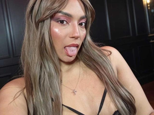 BrianaPratt tiene el pelo largo y ondulado, de color marrón, sombra de ojos rosa, un rostro brillante, y está sacando la lengua. Lleva un top negro con tirantes y un collar con una cruz..