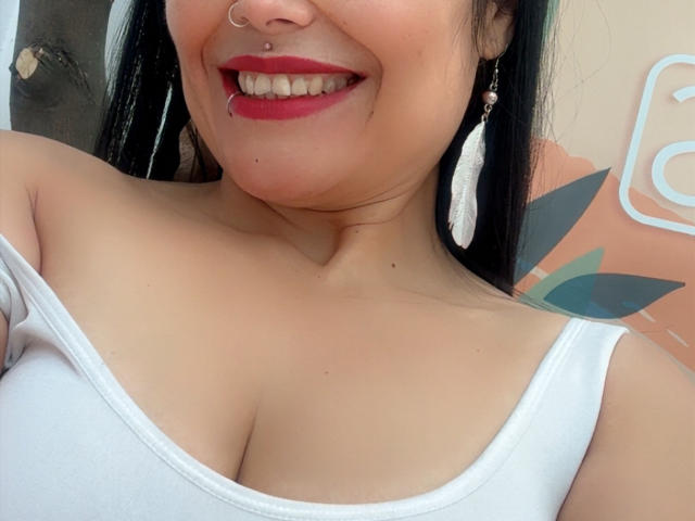 CharlotteCrofort's face with red lipstick, nose ring, and white feathers in ears, smiling, showing cleavage in a white tank top, her tits slightly visible, black hair, tan skin.