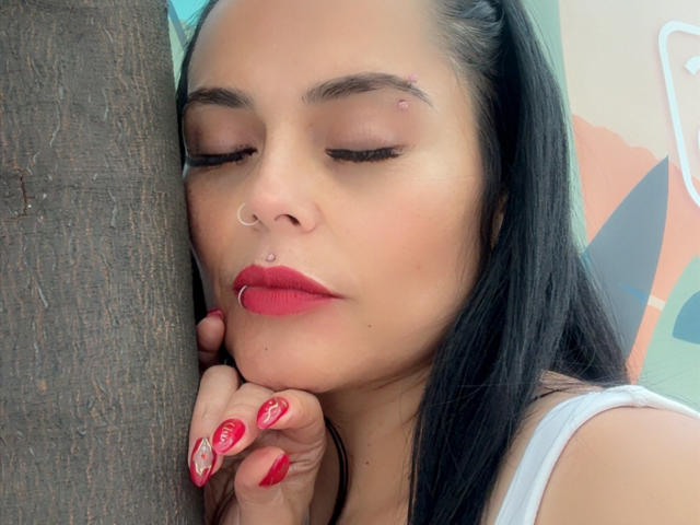 CharlotteCrofort with black hair, red lipstick, nose ring, and pink nails, eyes closed, resting head against dark wood, showing small acne scar on forehead.