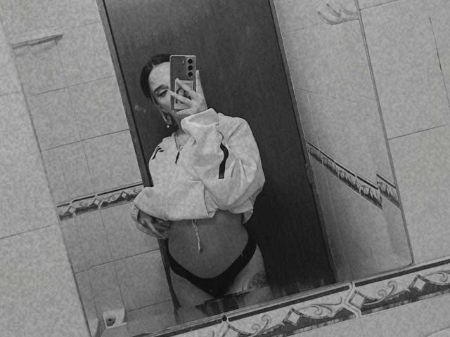 Autoportrait en noir et blanc d'AliceBellecourt, tenant un téléphone, portant une veste zippée et des sous-vêtements noirs, laissant apparaître son ventre et un tatouage sur sa hanche droite..
