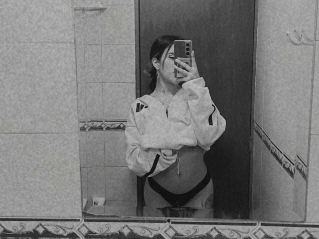 black and white mirror selfie of aliceBellecourt, showing her midriff, in a cropped jacket, black panties, with her hand covering her mouth, phone in the other hand.