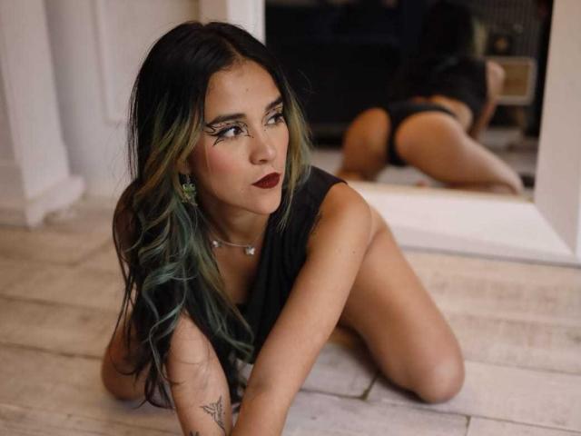 aliceBellecourt on wooden floor, black top, dark green and black hair, dark lipstick, kneeling with ass up, looking at mirror reflection, tattoo on right arm, butt visible.