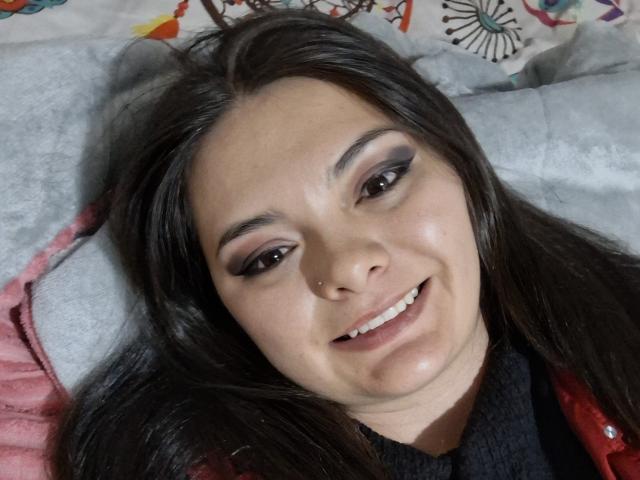 Katherin Latin hat langes, braunes Haar, eine kleine Nasenpiercing, dunklen Eyeliner und lächelt, wobei ihre Zähne zu sehen sind. Sie trägt eine rote Jacke über einem schwarzen T-Shirt..
